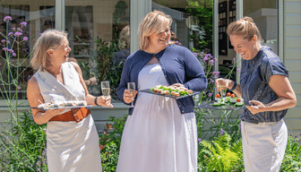 Three women carrying drinks and platters of food beside a garden room surrounded by colourful planting.